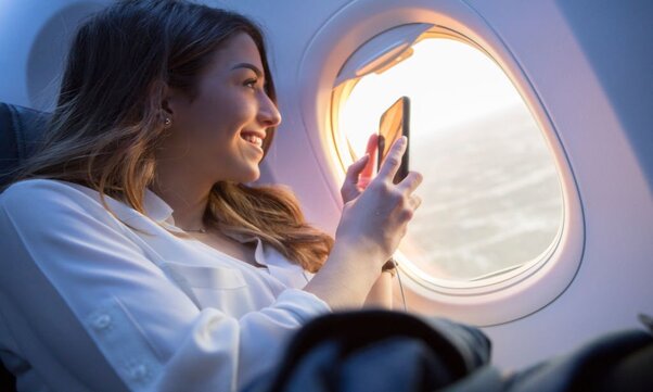 A smiling young woman sits in the window seat of a commercial airliner as it flies through the air.