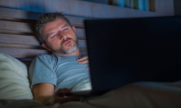 A man alone in bed using laptop talking to someone who uses an app to get paid to text.